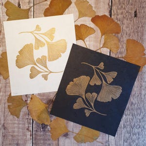 Op de afbeelding: Twee vierkante kunstprints met een botanische illustratie van gouden ginkgobladeren. De ene print heeft een witte achtergrond en de andere een zwarte achtergrond. Beide tonen dezelfde gedetailleerde tak met waaiervormige bladeren.