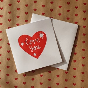 square white greetings card featuring a bright red heart with the words 'love you' and a star and sparkles in white.