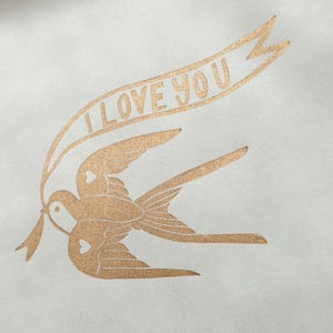 Op de afbeelding: Een gouden folie-afdruk van twee vogels die vliegen met een banner met de tekst "Ik hou van je".