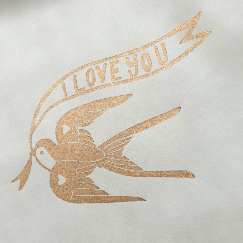 Op de afbeelding: Een gouden folie-afdruk van twee vogels die vliegen met een banner met de tekst "Ik hou van je".