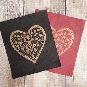 Op de afbeelding: Twee handgemaakte kaarten met een gouden hartontwerp op zwart en rood papier. Het hartontwerp heeft een bloemenpatroon.