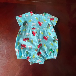 May include: A light blue baby onesie with a festive print of red cars, green trees, and white snowflakes.
