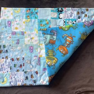 May include: A patchwork baby blanket with a blue and white color scheme. One side features a pattern of squares with various designs, including moons, fish, and bears. The other side has a blue background with cartoon monsters.