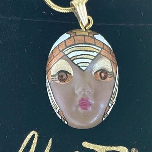 May include: A gold-colored necklace with a pendant featuring a painted face design. The pendant is brown with gold and white accents, and the necklace is paired with a black velvet pouch. The pouch has the word "Adagio" in gold lettering.
