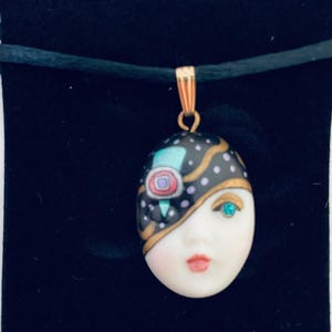 May include: A pendant featuring a painted face design with a gold-colored bail. The face has a blue gemstone eye, red lips, and a black and gold patterned mask. The pendant hangs on a black cord, displayed on a black background with the word "Rhapsody".