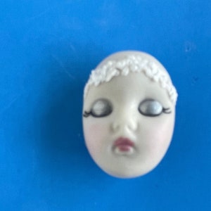 May include: A porcelain doll's head with closed eyes, painted eyelashes, and a small red mouth. The doll head has a white floral crown and a pale skin tone. The background is a solid blue.
