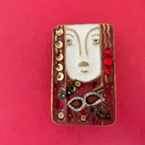 May include: Rectangular brooch featuring a stylized face with gold accents, red hair, and decorative elements. The face is white with gold details, set against a red background. Embellished with red and gold gems and a pair of brown and white glasses.