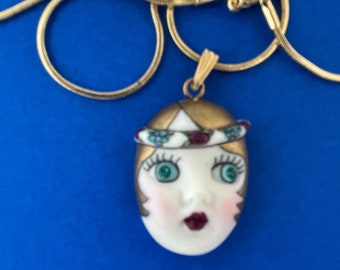 Op de afbeelding: Een goudkleurige ketting met een hanger met een geschilderd gezicht. De hanger heeft een wit gezicht met groene ogen, rode lippen en een goudkleurige hoofdbedekking met een decoratieve band. De ketting staat op een blauwe achtergrond.