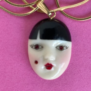 May include: A gold-tone necklace featuring a pendant shaped like a stylized face. The face has black hair, red lips, and red eyes. The pendant is attached to a thin, gold chain. The background is a solid pink color.