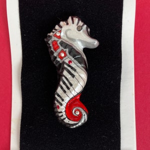 May include: A whimsical seahorse brooch with a piano key design. The seahorse is painted in silver, black, and red, with small red accents. The brooch is set against a black background.