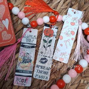 以下が含まれることがあります： さまざまなデザインとテキストが施された装飾的なブックマークのコレクション。あるブックマークには、本を読む骸骨と「I LIKE MY FAIRY-TALES DARK & DIRTY」と「BOOK WHORE」というテキストが描かれています。別のブックマークにはバラと「SPREAD THOSE PAGES」というテキストが描かれています。