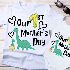 Op de afbeelding: Twee witte T-shirts met een groene dinosaurus en de tekst "Our 1st Mother's Day" erop.