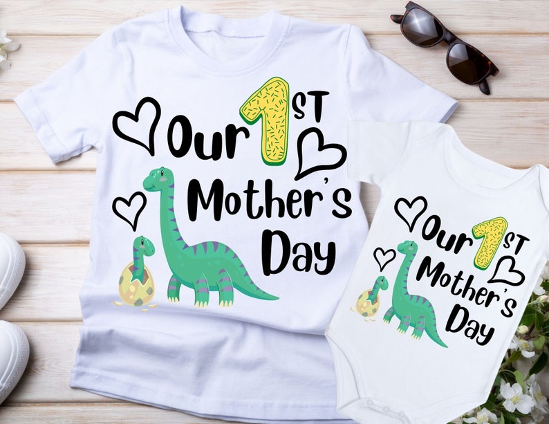 Op de afbeelding: Twee witte T-shirts met een groene dinosaurus en de tekst "Our 1st Mother's Day" erop.