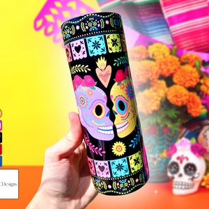 May include: A black and white tumbler with a colourful Day of the Dead design featuring two sugar skulls kissing. The skulls are decorated with flowers, hearts, and other festive details. The background is a black and white pattern.