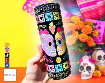 Op de afbeelding: Een zwarte en witte tumbler met een kleurrijk Dia de los Muertos-ontwerp met twee zoete schedels die elkaar kussen. De schedels zijn versierd met bloemen, harten en andere feestelijke details. De achtergrond is een zwart-wit patroon.
