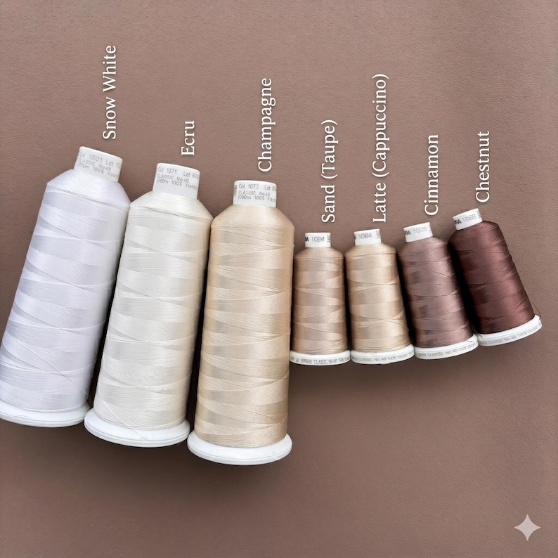 May include: Seven spools of sewing thread arranged in a row, ranging from light to dark shades. The colors are labeled as Snow White, Ecru, Champagne, Sand (Taupe), Latte (Cappuccino), Cinnamon, and Chestnut. Each spool has a white base.
