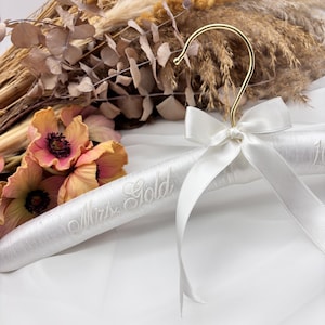 May include: A white padded clothes hanger featuring a gold-toned metal hook and a white satin ribbon bow tied at the neck. The hanger is embroidered with the text Mrs. Gold in a cursive script font, providing a decorative display for garments.