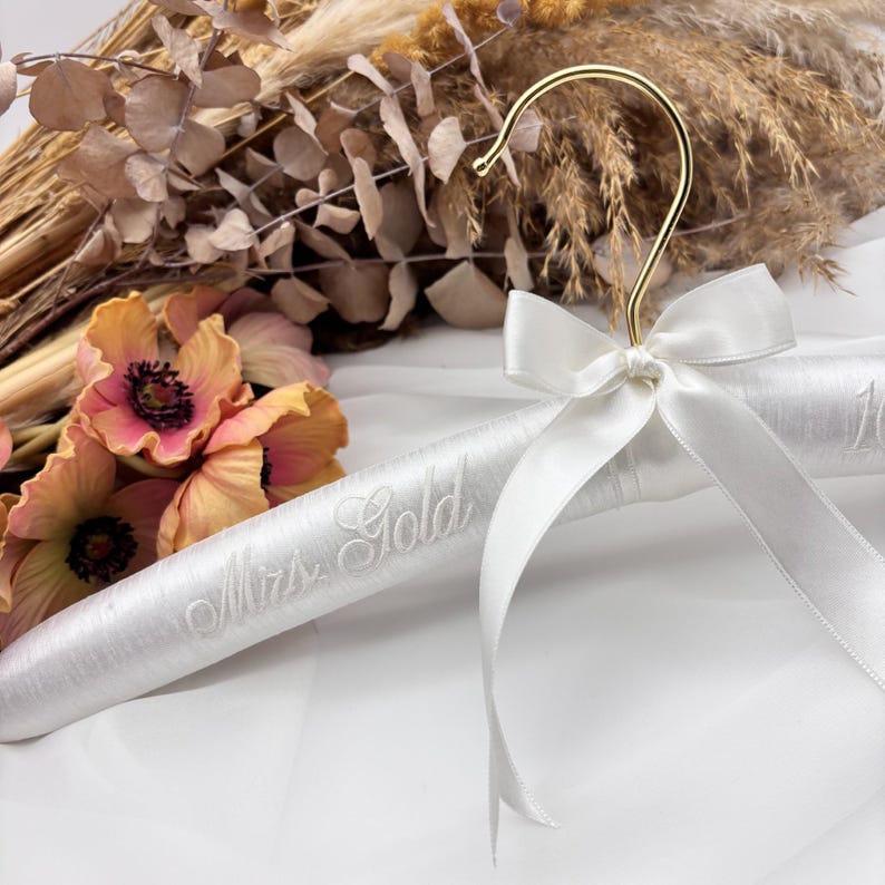 May include: A white padded clothes hanger featuring a gold-toned metal hook and a white satin ribbon bow tied at the neck. The hanger is embroidered with the text Mrs. Gold in a cursive script font, providing a decorative display for garments.