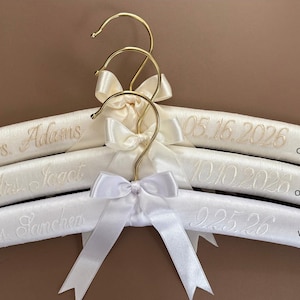 May include: Three padded hangers with gold-tone metal hooks and satin bows. The top hanger is cream fabric with 'Mrs. Adams' and '05.16.2026' embroidered. The middle is off-white with 'Mrs. Isaac' and '10.10.2028'. The bottom is white with 'Mrs. Sanchez' and '9.25.26'.