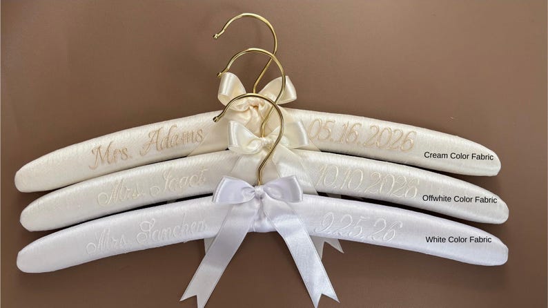 May include: Three padded hangers with gold-tone metal hooks and satin bows. The top hanger is cream fabric with 'Mrs. Adams' and '05.16.2026' embroidered. The middle is off-white with 'Mrs. Isaac' and '10.10.2028'. The bottom is white with 'Mrs. Sanchez' and '9.25.26'.