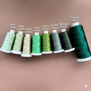 May include: Eight spools of sewing thread arranged in a row, displaying a gradient of green shades labeled Mint Green, Sage Green, Olive Green, Kelly Green, Moss Green, Hunter Green, Midnight Green, and Emerald Green on white labels.