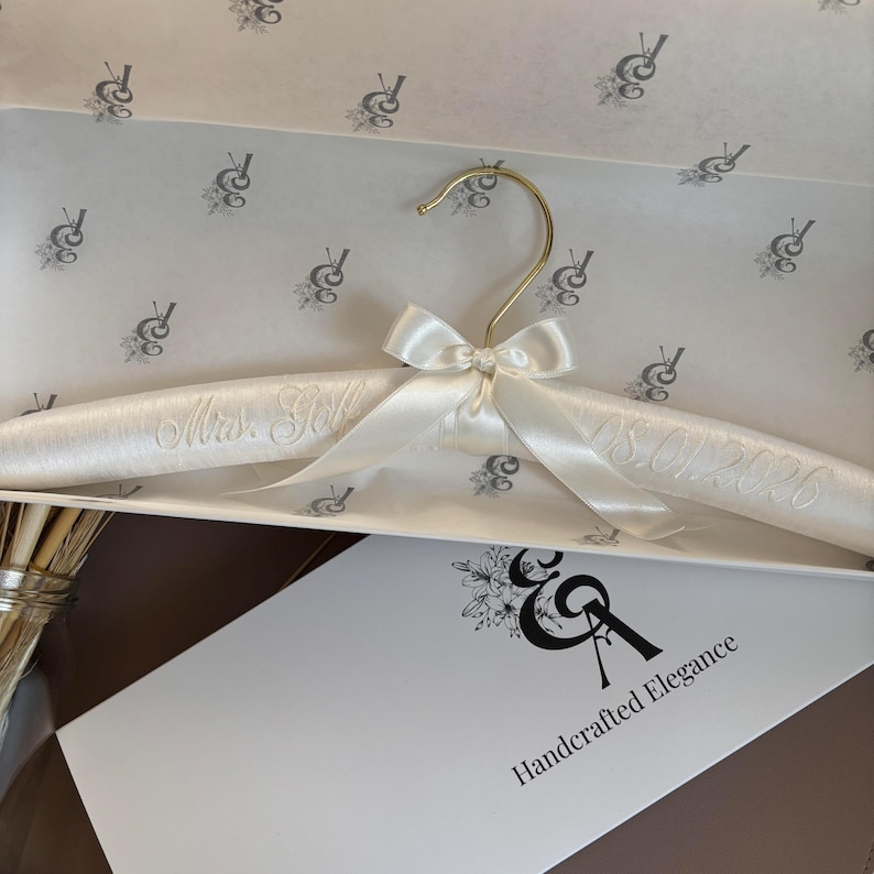 May include: A padded ivory satin hanger featuring a gold-tone metal hook and a matching satin bow at the center. The hanger is embroidered with the text Mrs. Gobl and the date 08.01.2026. It is presented inside a white box with a floral logo.