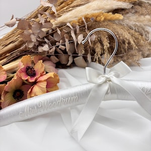 May include: A white satin-covered clothes hanger featuring a silver metal hook and a white satin ribbon bow tied at the neck. The hanger is embroidered with the text Mrs. Woodrum in a cursive script font across the front of the padded surface.