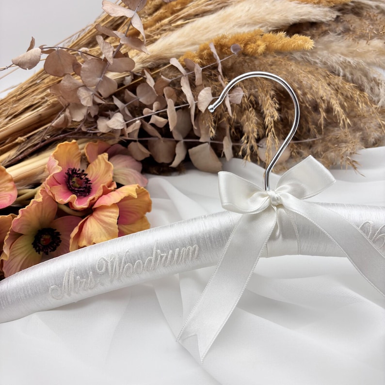 May include: A white satin-covered clothes hanger featuring a silver metal hook and a white satin ribbon bow tied at the neck. The hanger is embroidered with the text Mrs. Woodrum in a cursive script font across the front of the padded surface.