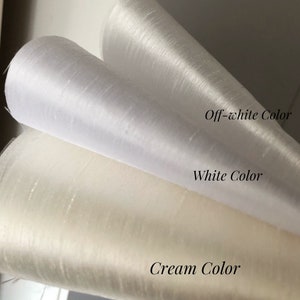 May include: Three rolls of fabric with a subtle sheen and textured surface. The top roll is labeled Off-white Color, the middle roll is labeled White Color, and the bottom roll is labeled Cream Color, showcasing distinct shades of light fabric.
