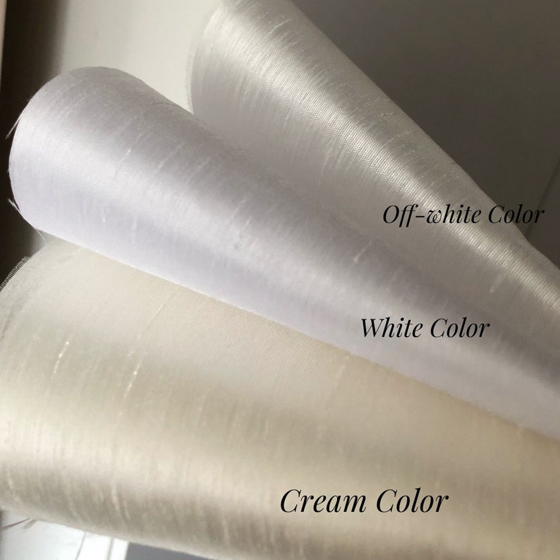 May include: Three rolls of fabric with a subtle sheen and textured surface. The top roll is labeled Off-white Color, the middle roll is labeled White Color, and the bottom roll is labeled Cream Color, showcasing distinct shades of light fabric.