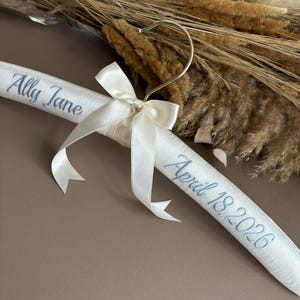 May include: A padded white satin clothes hanger featuring a gold metal hook and a decorative cream-colored ribbon bow. The hanger is embroidered with the name Ally Jane on the left side and the date April 18, 2026, on the right side in gray.