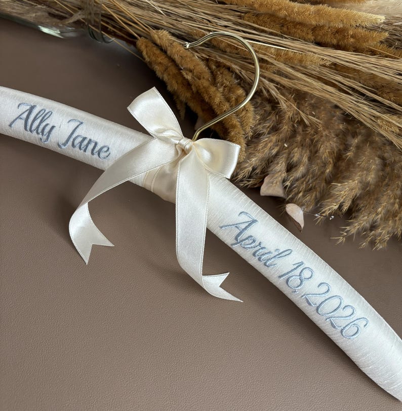 May include: A padded white satin clothes hanger featuring a gold metal hook and a decorative cream-colored ribbon bow. The hanger is embroidered with the name Ally Jane on the left side and the date April 18, 2026, on the right side in gray.