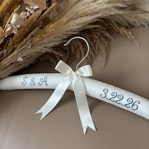 May include: A white padded clothes hanger featuring a silver metal hook and a white satin ribbon bow. The hanger is embroidered with the blue text S & A on the left side and the date 3.22.26 on the right side in a cursive script font.
