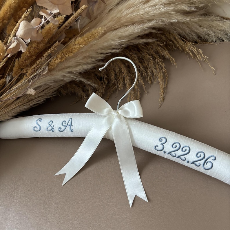 May include: A white padded clothes hanger featuring a silver metal hook and a white satin ribbon bow. The hanger is embroidered with the blue text S & A on the left side and the date 3.22.26 on the right side in a cursive script font.
