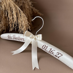 May include: A padded white satin clothes hanger featuring a silver metal hook and a central cream-colored ribbon bow. The hanger is embroidered with the brown text Mrs. Blackburn on the left side and the date 01.16.27 on the right side.