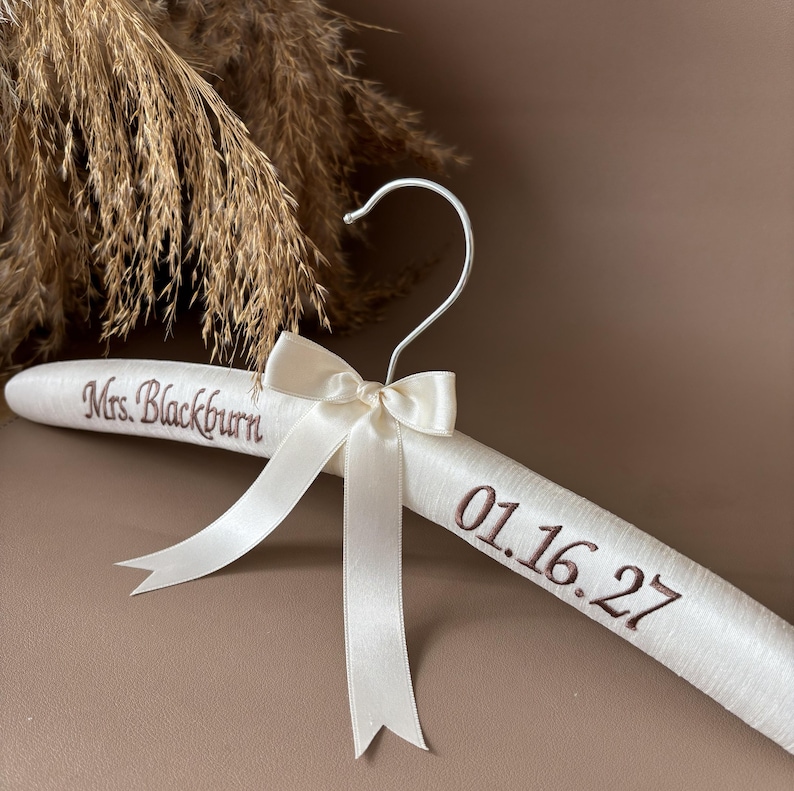 May include: A padded white satin clothes hanger featuring a silver metal hook and a central cream-colored ribbon bow. The hanger is embroidered with the brown text Mrs. Blackburn on the left side and the date 01.16.27 on the right side.
