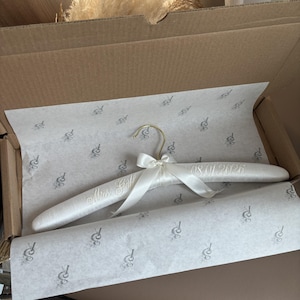 May include: A padded white satin clothes hanger featuring a gold-toned metal hook and a white satin ribbon bow tied at the center. The hanger is embroidered with the text Mrs Gohl on the left side and the date 08.01.2026 on the right side.