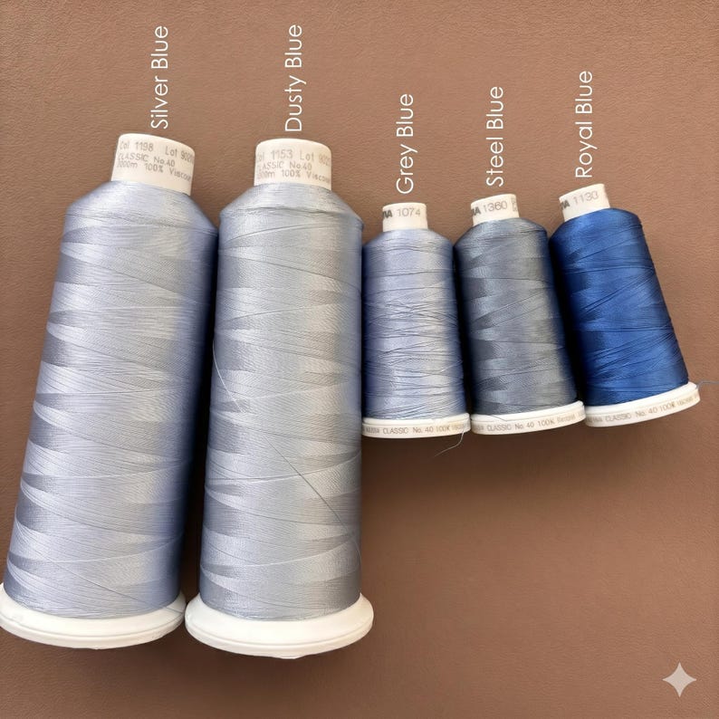 May include: Five spools of embroidery thread in varying shades of blue, labeled Silver Blue, Dusty Blue, Grey Blue, Steel Blue, and Royal Blue. Each spool features a white base and top, with the thread color name printed above each individual spool.