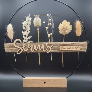 Könnte beinhalten: Ein rundes Holzschild mit dem Namen "Serums" in Kursivschrift auf einem Jute-Hintergrund. Das Schild ist umgeben von getrockneten Blumen und Gräsern, darunter eine weiße Margerite, eine Mohnkapsel und ein Pampasgras. Das Schild hängt an einem schwarzen Metallring und hat einen kleinen Holzsockel mit einem eingegrabenen Herzen. Das Schild enthält auch den Text "Schie, dass du bist!"