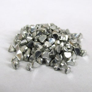 May include: A pile of small, silver-colored metal pieces, each with multiple facets and a semi-circular shape. The pieces are scattered on a white surface, creating a textured appearance. The metallic objects appear to be industrial components.