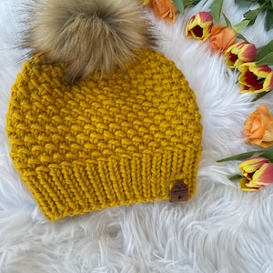 Può includere: Berretto lavorato a maglia giallo senape con un grande e soffice pompon marrone chiaro sulla parte superiore. Il cappello ha un motivo a punto strutturato e una piccola etichetta in pelle marrone. Il berretto è esposto su una superficie bianca e soffice, con tulipani arancioni e gialli.