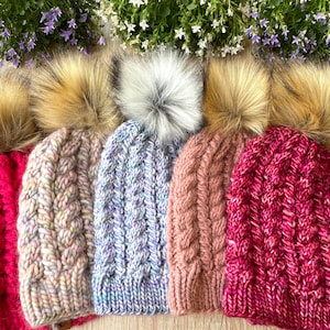 Può includere: Cinque berretti a trecce nei toni del rosso, rosa e blu, ciascuno sormontato da un pompon in finta pelliccia. I cappelli sono disposti in fila, mostrando i loro motivi strutturati e i vari colori. Accenti floreali visibili sullo sfondo.
