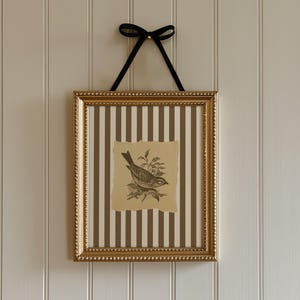 Puede incluir: Una obra de arte enmarcada con una ilustración de un pájaro. El marco dorado contiene un dibujo de un pájaro sobre papel, sobre un fondo a rayas marrones y blancas. Se utiliza una cinta negra para colgarlo.