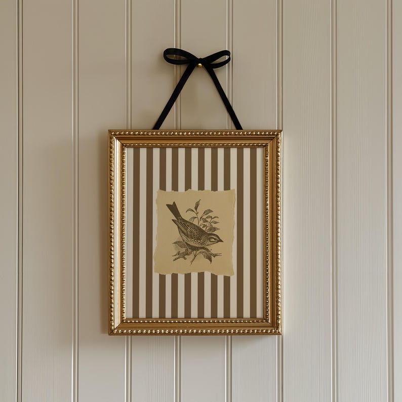 Op de afbeelding: Een ingelijst kunstwerk met een vogelillustratie. De goudkleurige lijst bevat een papier met een vogeltekening, tegen een gestreepte achtergrond van bruin en wit. Een zwart lint wordt gebruikt om op te hangen.
