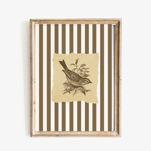 Op de afbeelding: Een ingelijst kunstwerk met een gedetailleerde illustratie van een vogel op een tak. De vogel staat tegen een gescheurd, bruin papier, dat gecentreerd is op een gestreepte achtergrond van bruine en cr&egrave;mekleurige verticale lijnen. De lijst heeft een lichte houtkleur.