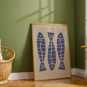 Op de afbeelding: Een ingelijst print met een minimalistisch ontwerp met drie blauwe vissen silhouetten op een cr&egrave;me achtergrond. De lijst is gemaakt van hout en heeft een natuurlijke afwerking.