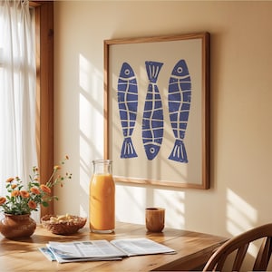 Op de afbeelding: Een ingelijst afdruk van drie blauwe vissen met witte contouren op een witte achtergrond. De afdruk hangt aan een beige muur in een kamer met een houten tafel en een glazen kan met sinaasappelsap.