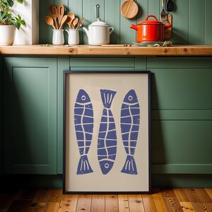 Op de afbeelding: Een ingelijst afdruk van drie blauwe vissen met witte contouren op een cr&egrave;mekleurige achtergrond. De afdruk hangt aan een groene muur in een keuken.