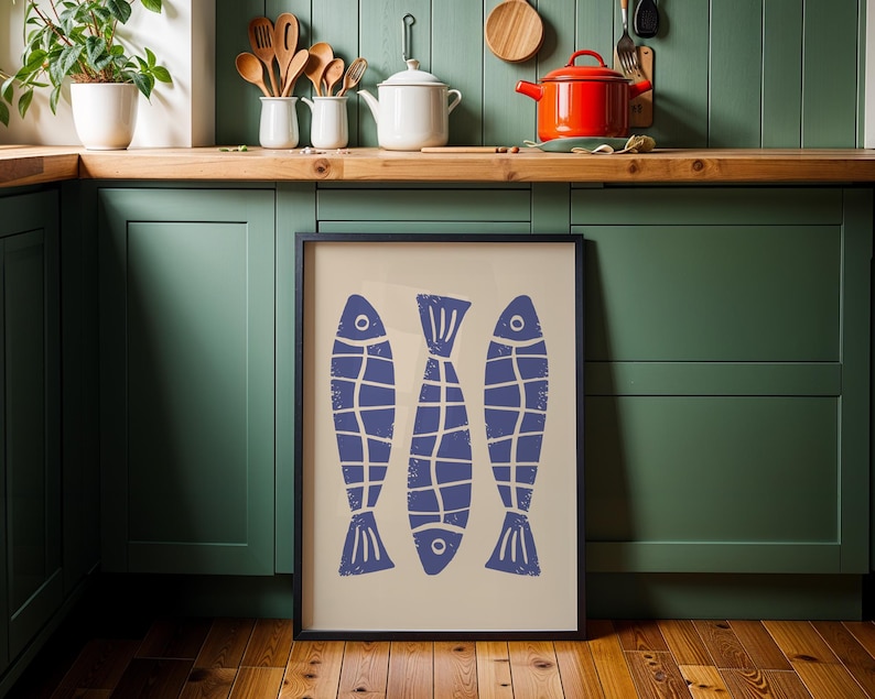 Op de afbeelding: Een ingelijst afdruk van drie blauwe vissen met witte contouren op een cr&egrave;mekleurige achtergrond. De afdruk hangt aan een groene muur in een keuken.