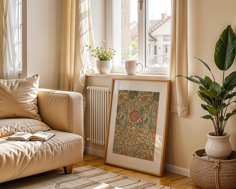 May include: A beige sofa with a book on it, a framed print with a floral design, a potted plant, and a window with sunlight streaming in.